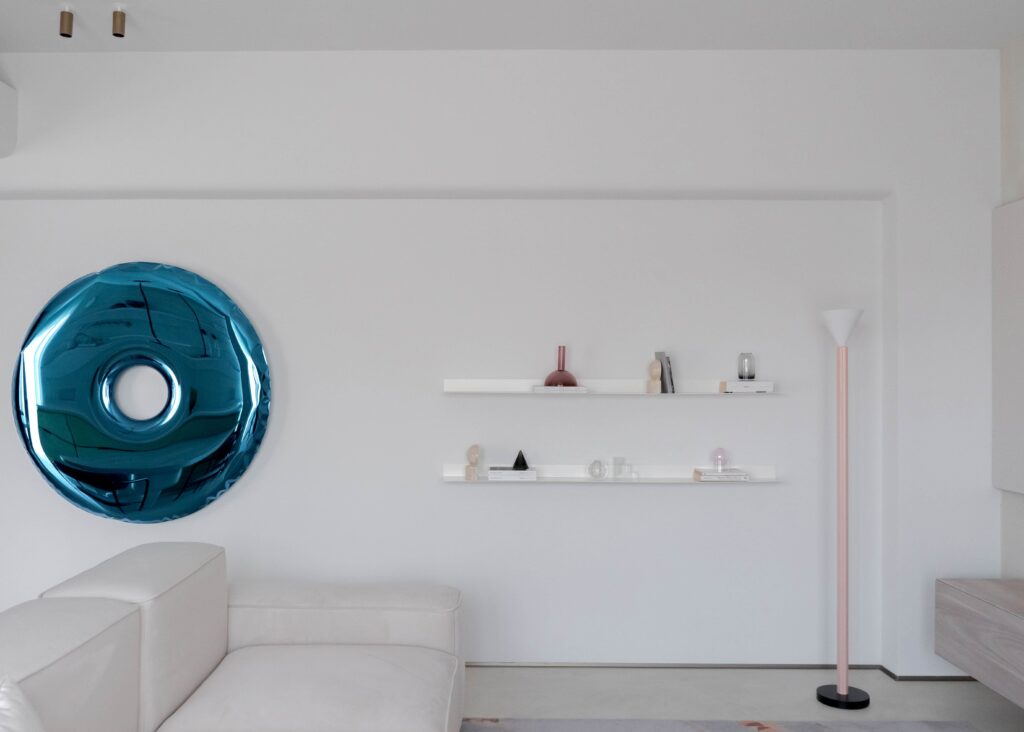 Living minimalist văzut frontal, cu oglindă decorativă albastră, rafturi albe și lampadar roz