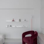 Living cu fotoliu burgundy Cassina Dudet, rafturi albe și accente pastelate într-un apartament minimalist glam